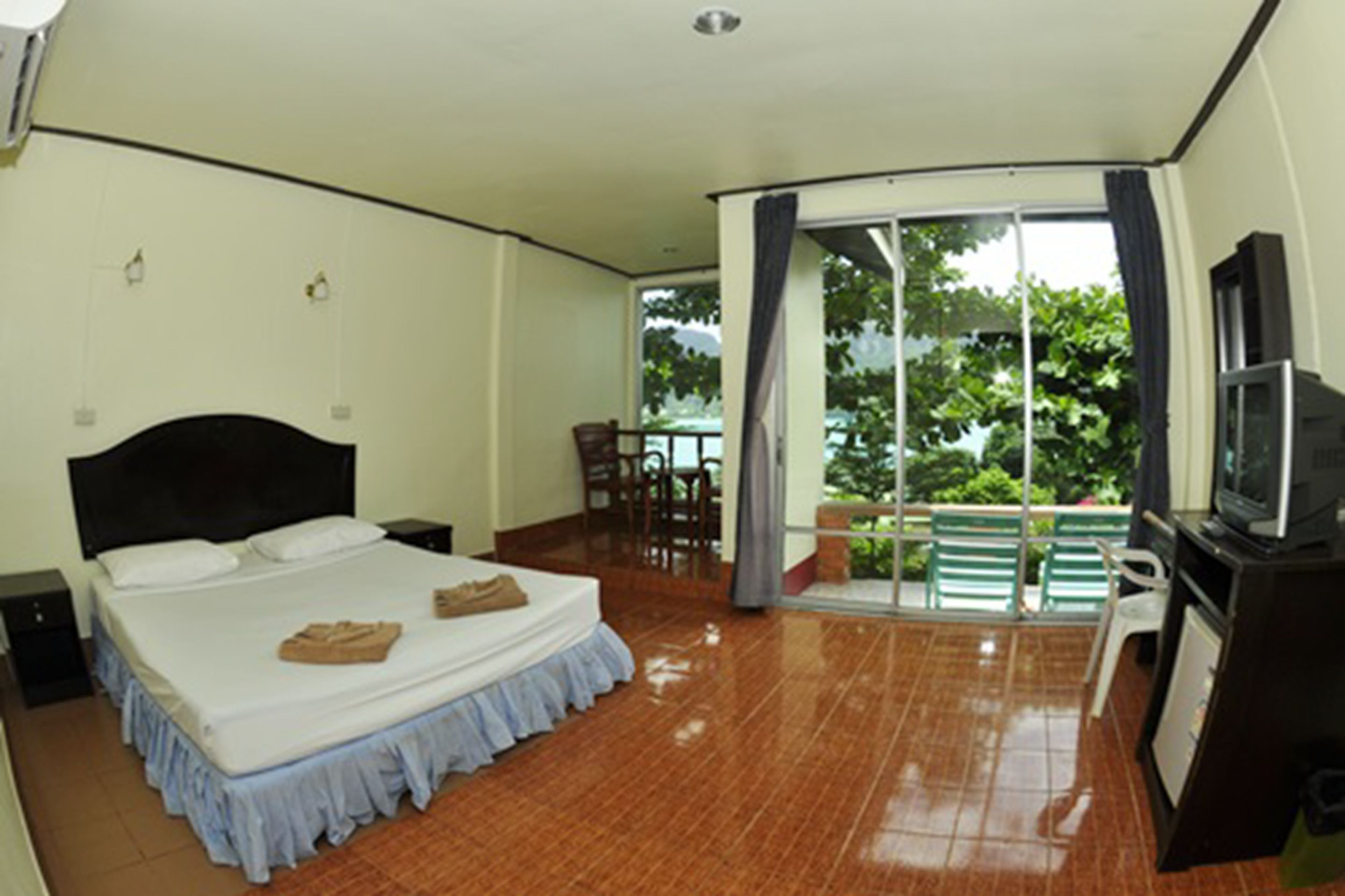 A hotel room with a bed, a TV, a balcony with chairs, and a view of greenery outside.