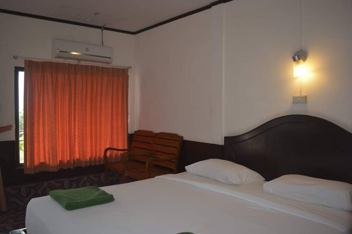 A hotel room with a bed, a chair, an air conditioning unit, a window with curtains, and a wall-mounted light.