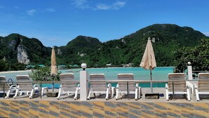 A poolside area with lounge chairs and umbrellas, overlooking a turquoise sea and lush green mountains.