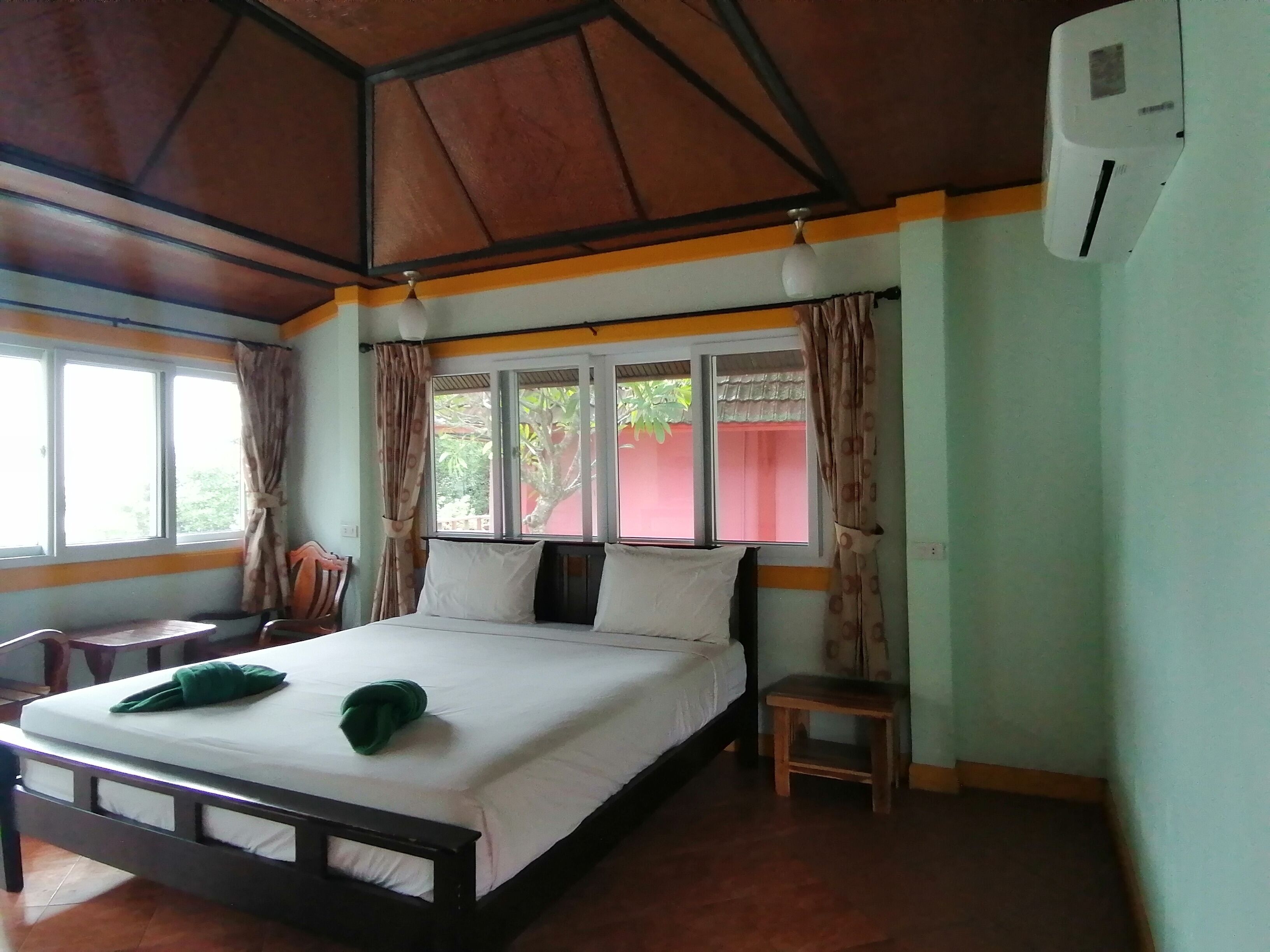 A hotel room with a bed, a small table, a chair, a window with curtains, and an air conditioning unit.