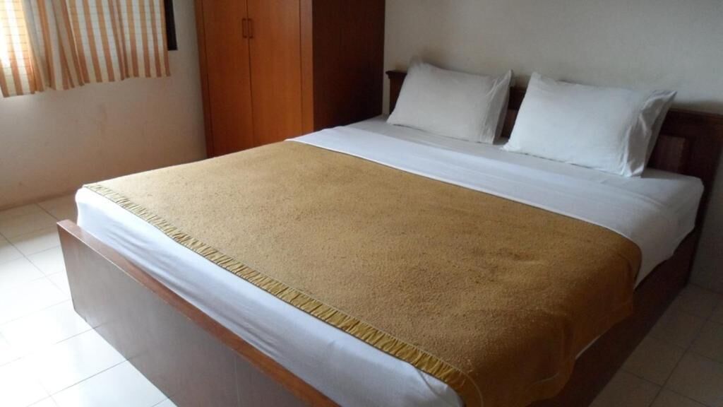 Un lit simple avec un matelas blanc et une couverture marron, une armoire en bois et un mur uni avec une fenêtre.