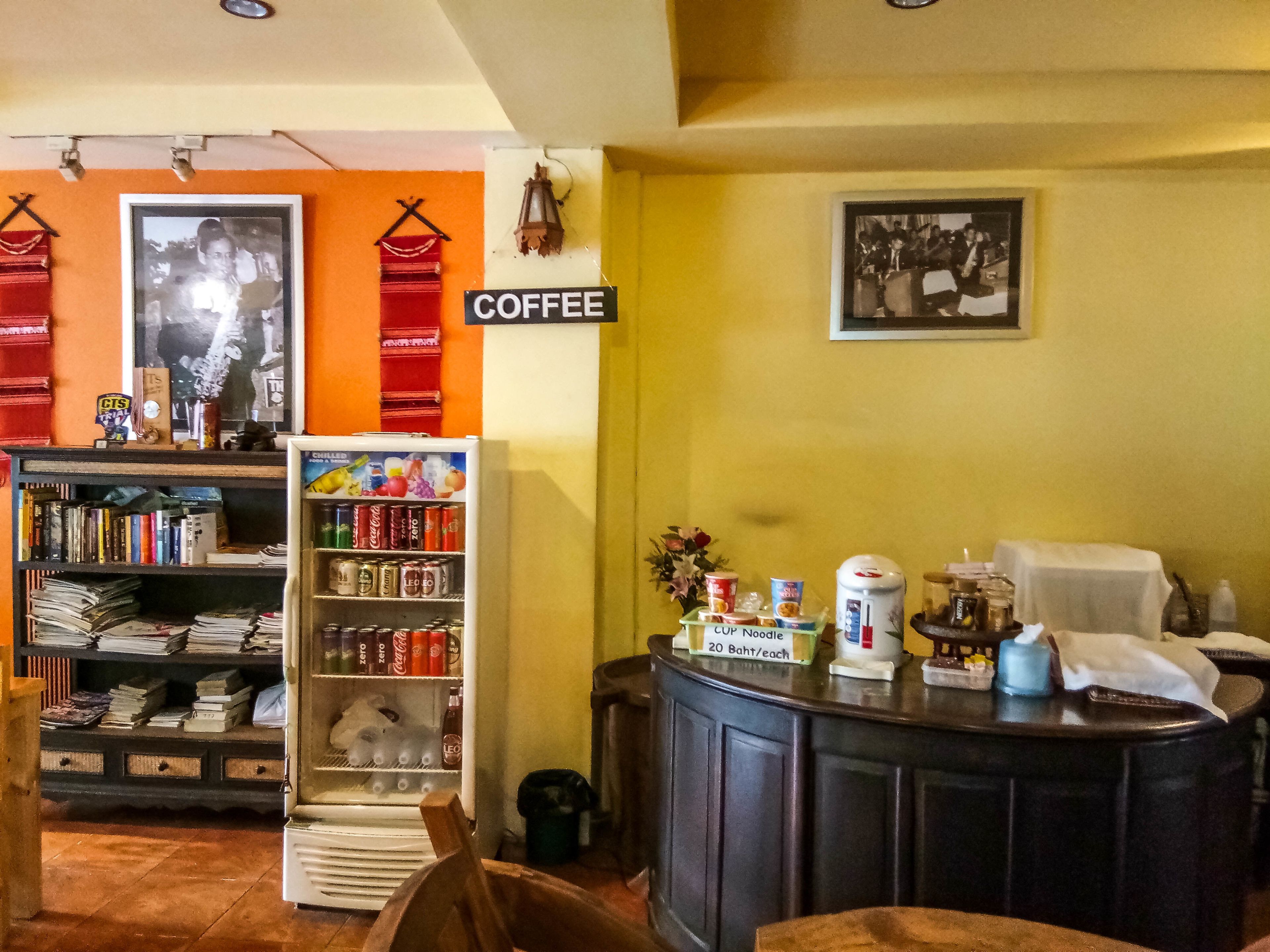 Interior acogedor de una cafetería con un letrero de café, una estantería, un refrigerador lleno de bebidas y una barra con varios artículos.