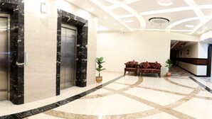 Um hall com piso de mármore, dois elevadores, sofás com padrão vermelho e plantas em vasos.