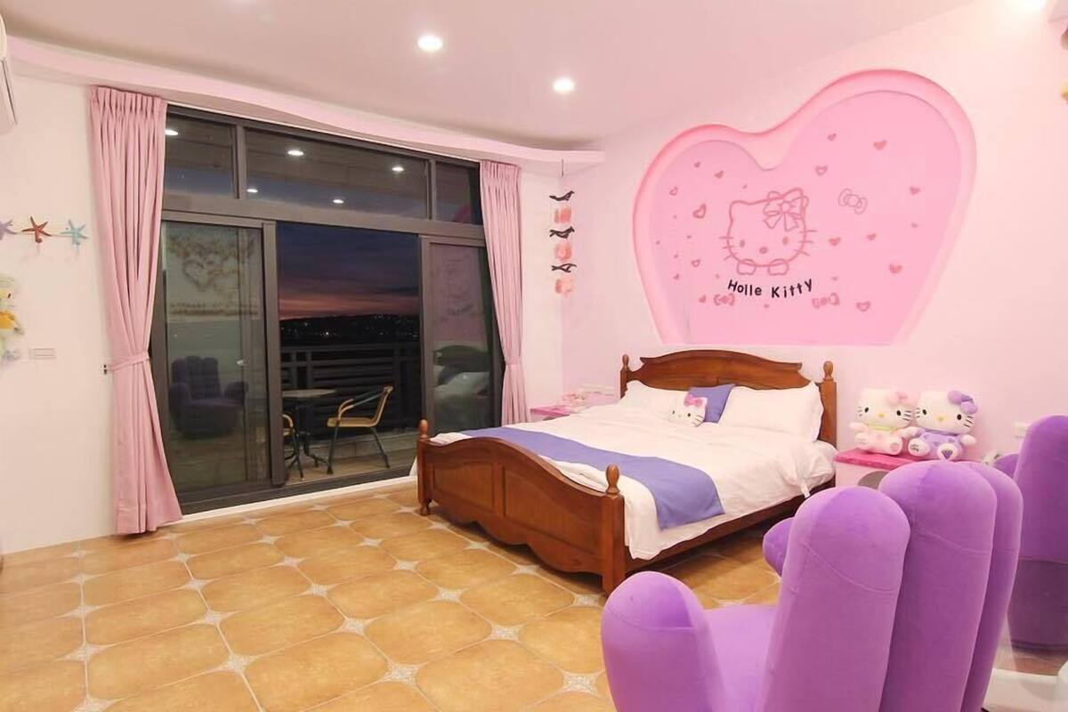 A bedroom with a large window, a bed with a wooden headboard, a pink wall decoration, and purple chairs.