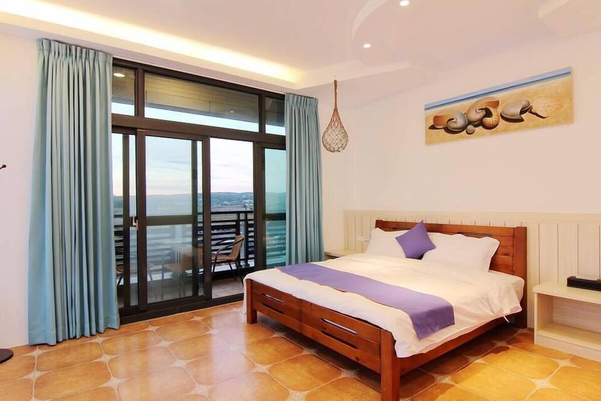 A bedroom with a large bed, a balcony with a view, and a hanging light fixture.