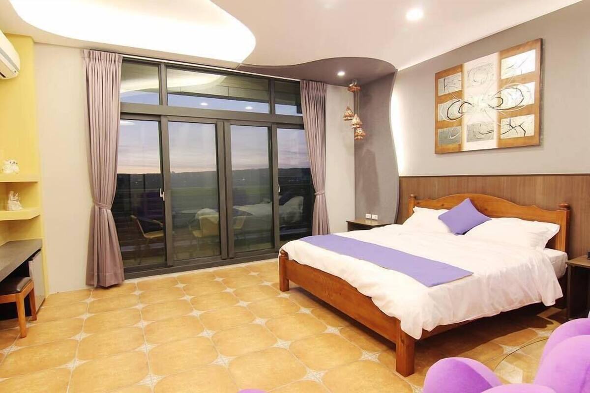 A bedroom with a large bed, a desk, and a view through a sliding glass door.