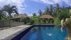 A swimming pool with a statue, surrounded by palm trees and buildings with tiled roofs.