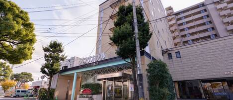The exterior of Plaza Hotel Fujino with a modern facade, a covered entrance, and a parking area visible in the foreground.