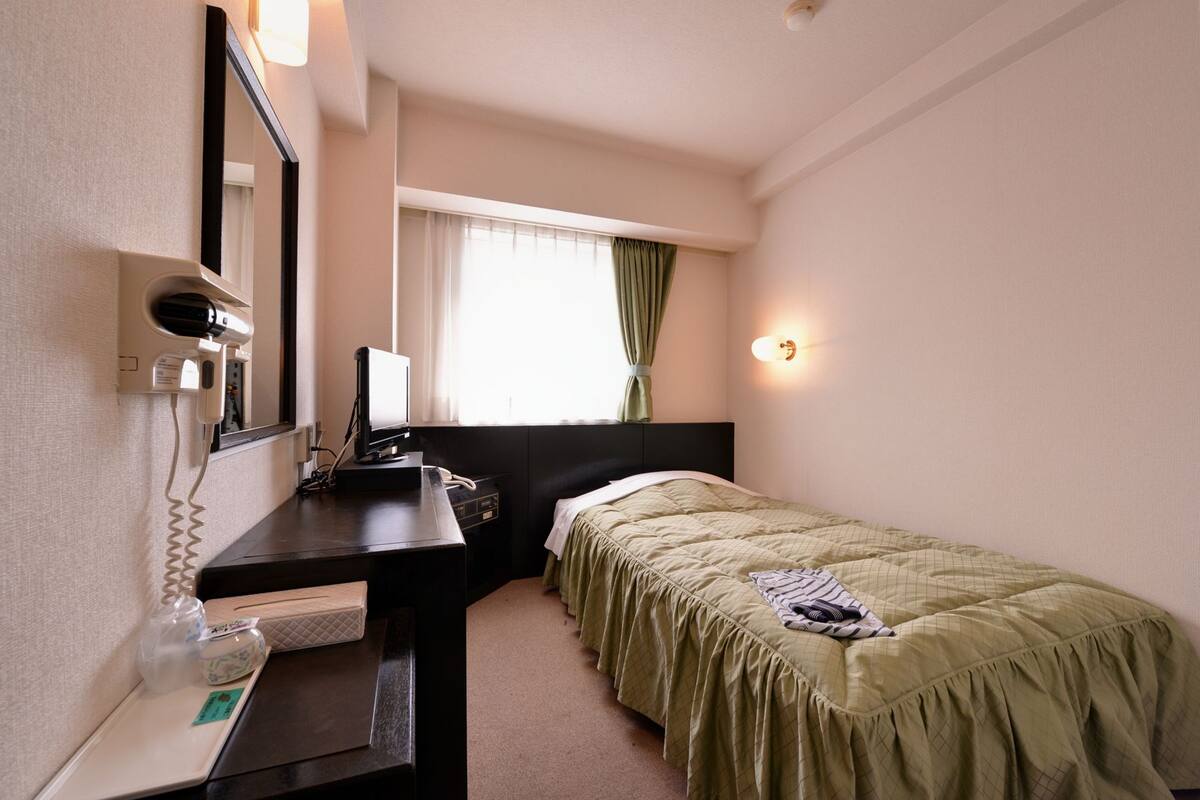 Habitación de hotel con una cama, un escritorio con computadora, un espejo y una ventana con cortinas.
