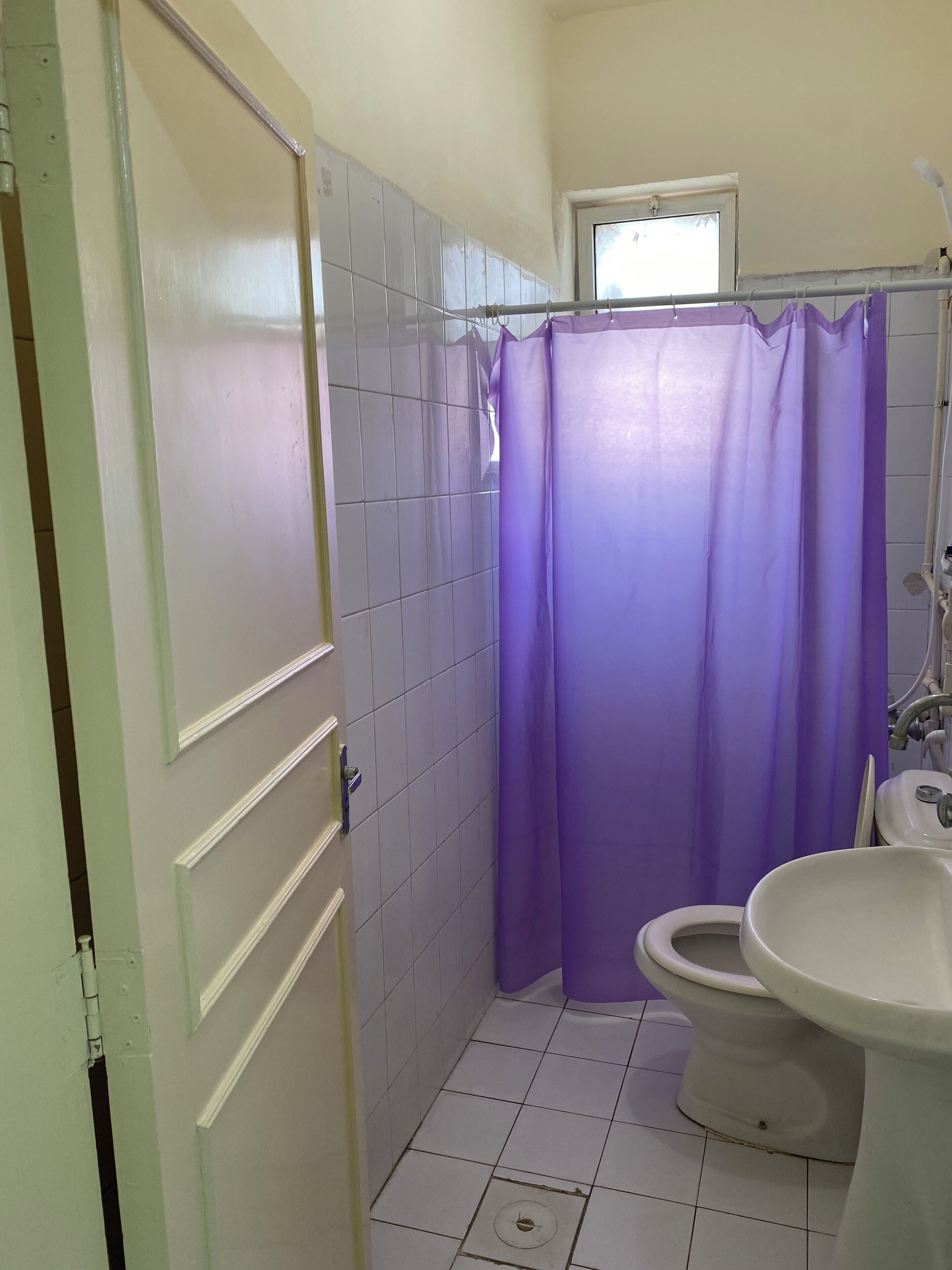 Een badkamer met witte betegelde wanden, een paars douchegordijn, een wit toilet en een witte wastafel.