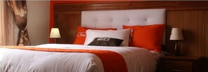 Ein Hotelzimmer mit einem großen Bett, orange-weißer Bettwäsche, einem Holzkopfteil und zwei Nachttischlampen.