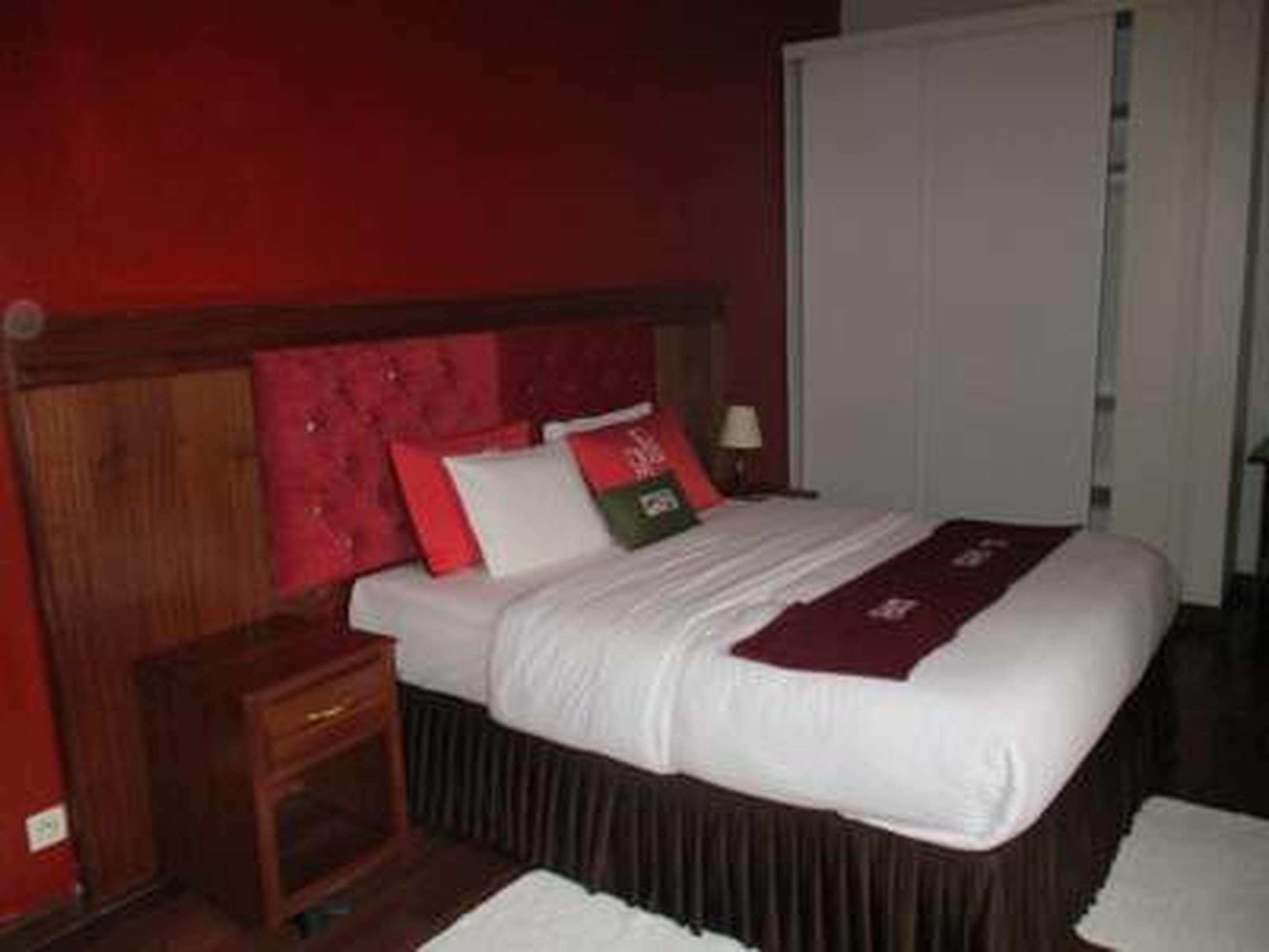 A hotel room with a wooden headboard, a bed with white bedding and red and green pillows, a nightstand, and a white door.