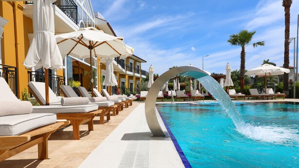 Un área junto a la piscina con sillones de descanso y sombrillas, una fuente de agua y un edificio con balcones.