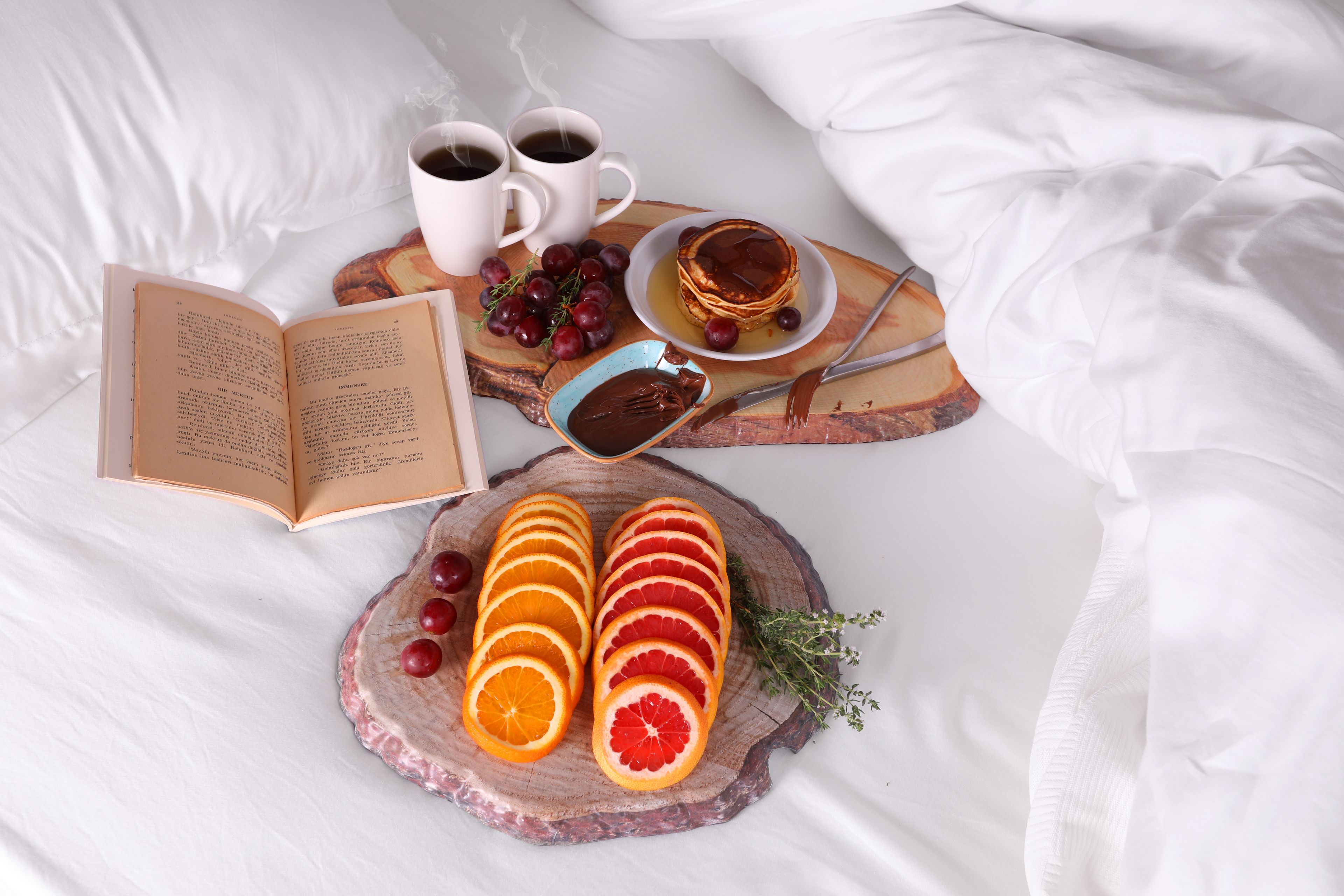 Un desayuno ordenado sobre la cama, con un libro, dos tazas de café, una pila de panqueques, un cuenco de chocolate, naranjas en rodajas y uvas.