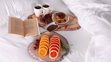 Un desayuno ordenado sobre la cama, con un libro, dos tazas de café, una pila de panqueques, un cuenco de chocolate, naranjas en rodajas y uvas.