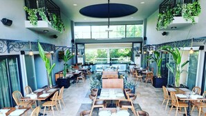 Un restaurante espacioso con varias mesas y sillas, ventanas amplias y plantas de interior.