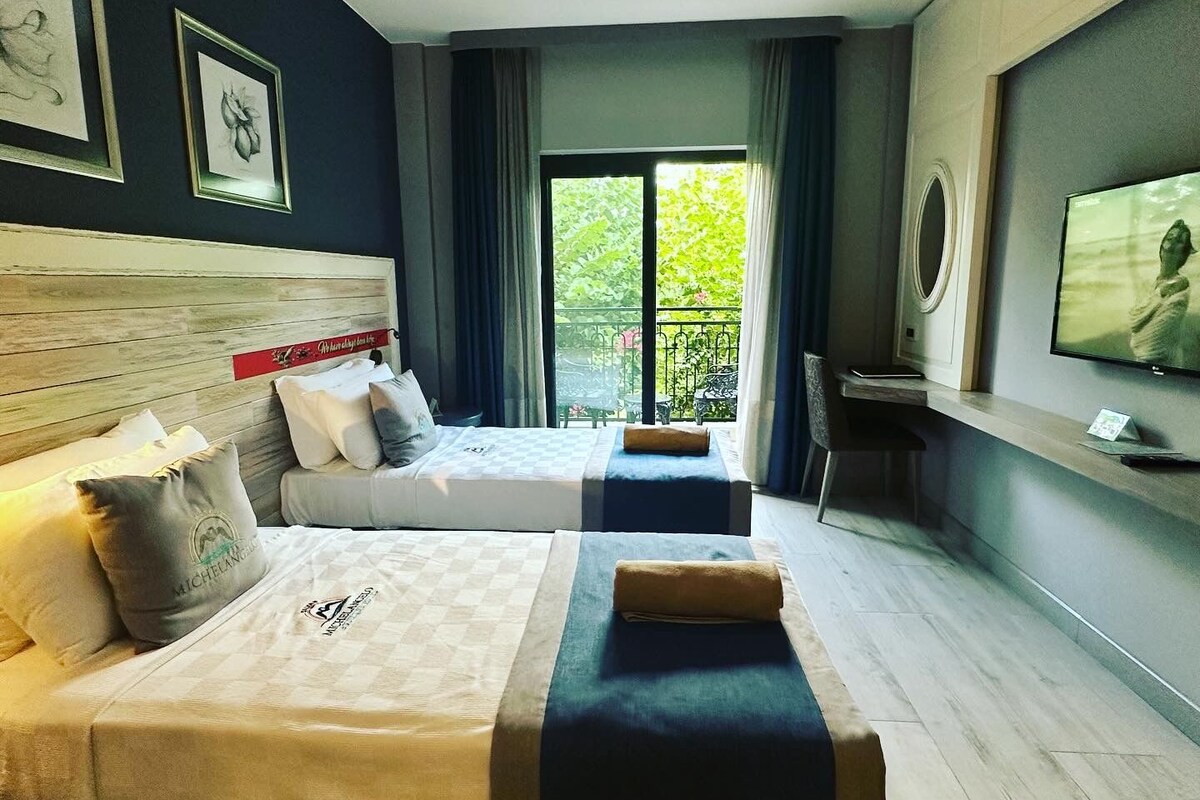 A hotel room with two beds, a television, and a balcony with a view of greenery.