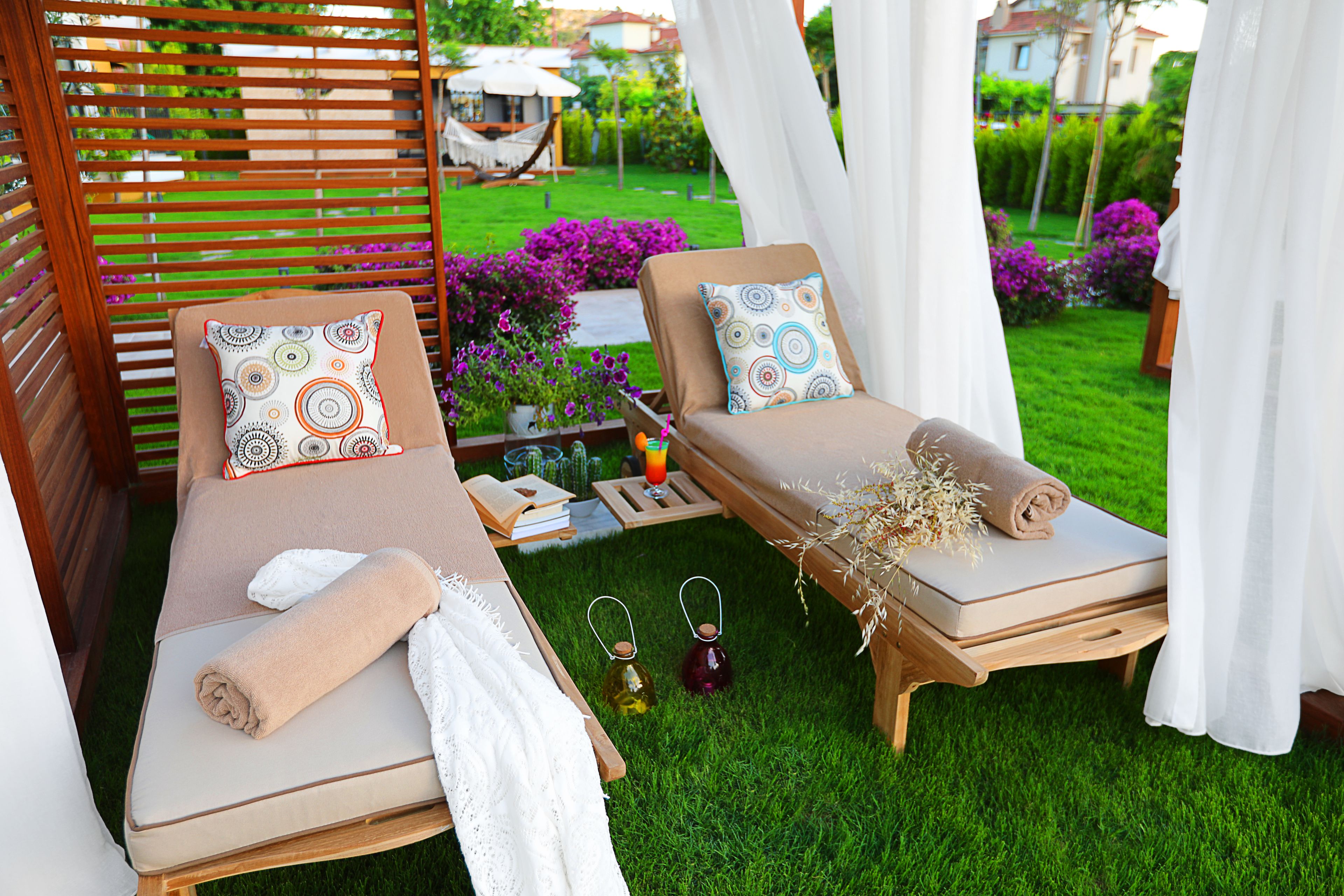 Dos tumbonas con toallas y almohadones decorativos, una mesita con un libro y una bebida, y un exuberante jardín al fondo.