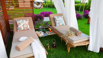 Dos tumbonas con toallas y almohadones decorativos, una mesita con un libro y una bebida, y un exuberante jardín al fondo.