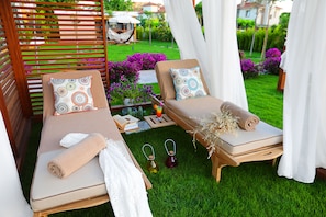 Two sunbeds with towels and decorative pillows, a small table with a book and a drink, and a lush green garden in the background.