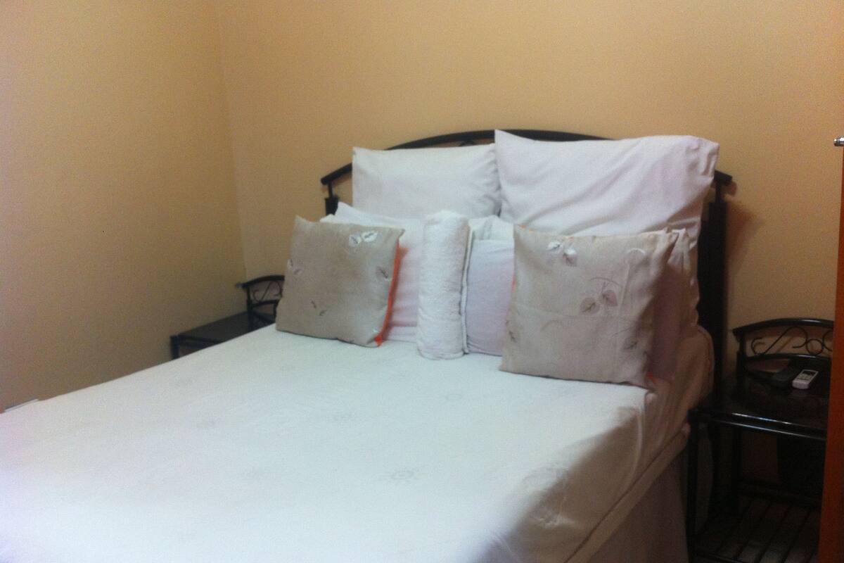 A neatly made bed with white linens and pillows, a black metal headboard, and a small bedside table with a lamp.
