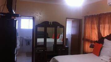 A bedroom with a bed, a dresser, a mirror, a wardrobe, a window, and a bathroom with a toilet and sink.
