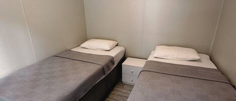 A small, single-room accommodation with two single beds, each with a pillow and a blanket.