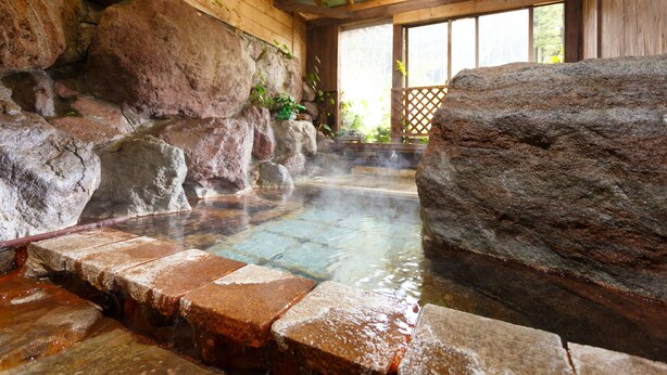 Un área de aguas termales con escalones de piedra que bajan al agua, rodeada de grandes rocas y paredes de madera.