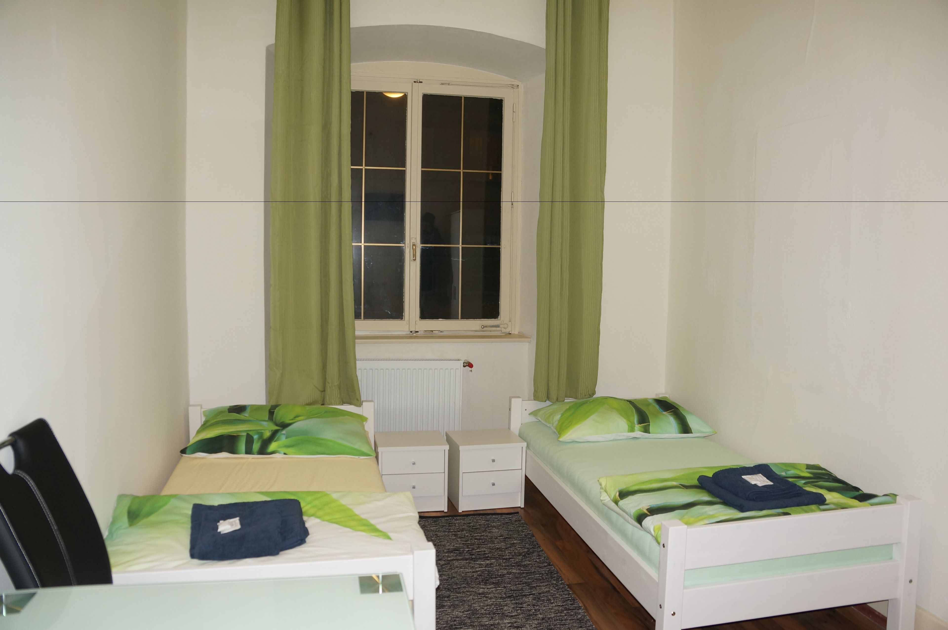 Une chambre avec deux lits, une fenêtre avec des rideaux verts, un radiateur et une petite table.