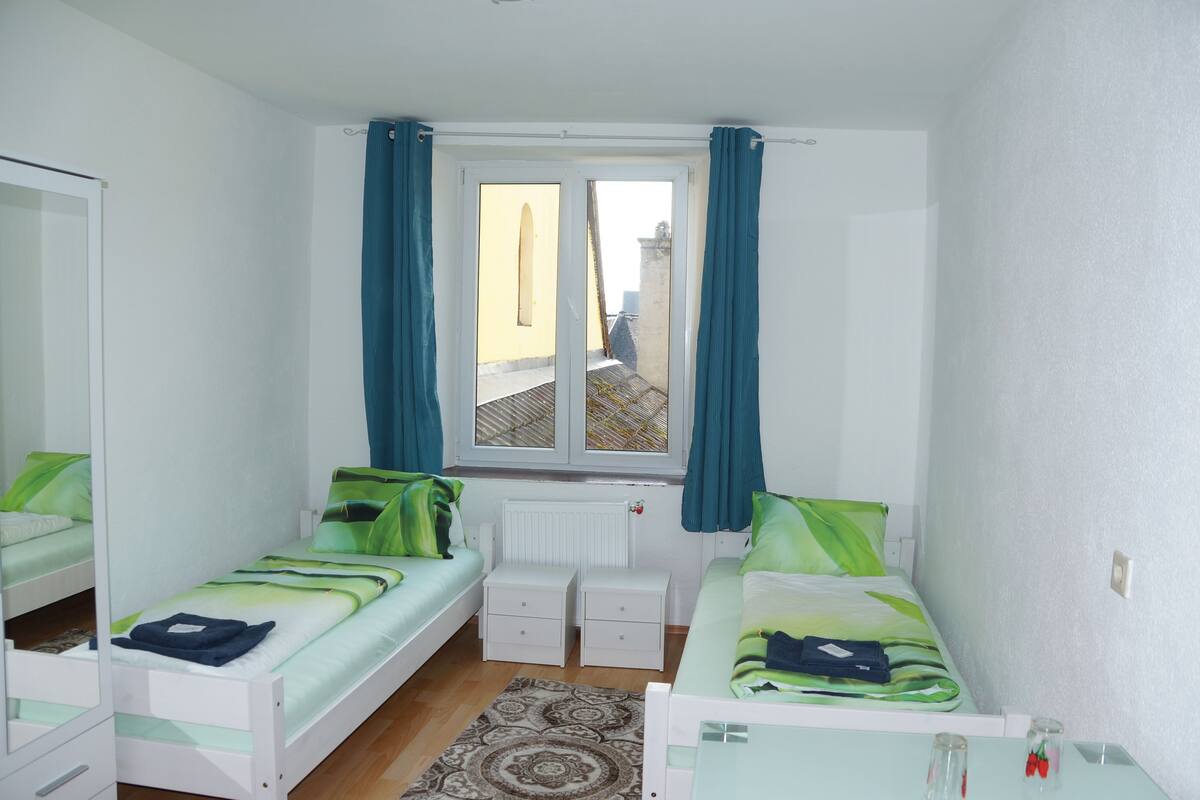 A small, well-lit bedroom with two single beds, a window with curtains, a radiator, and a mirror.
