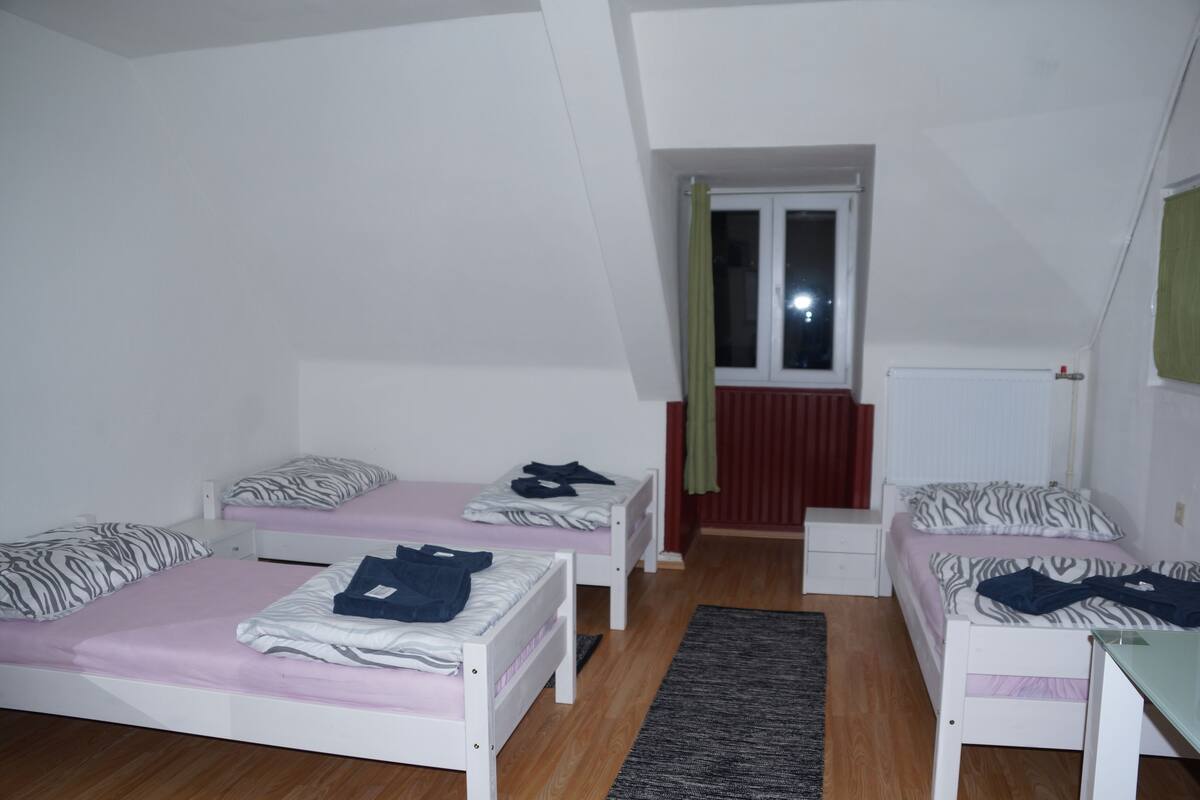A small, well-lit room with two single beds, a small table, and a window with curtains.