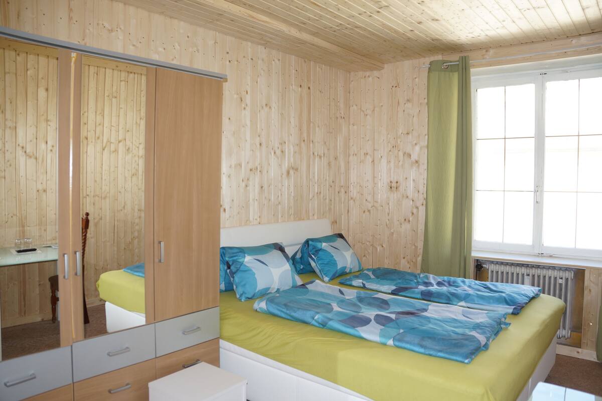 A room with wooden panel walls, a bed with blue and green bedding, a mirror cabinet, and a window with a green curtain.
