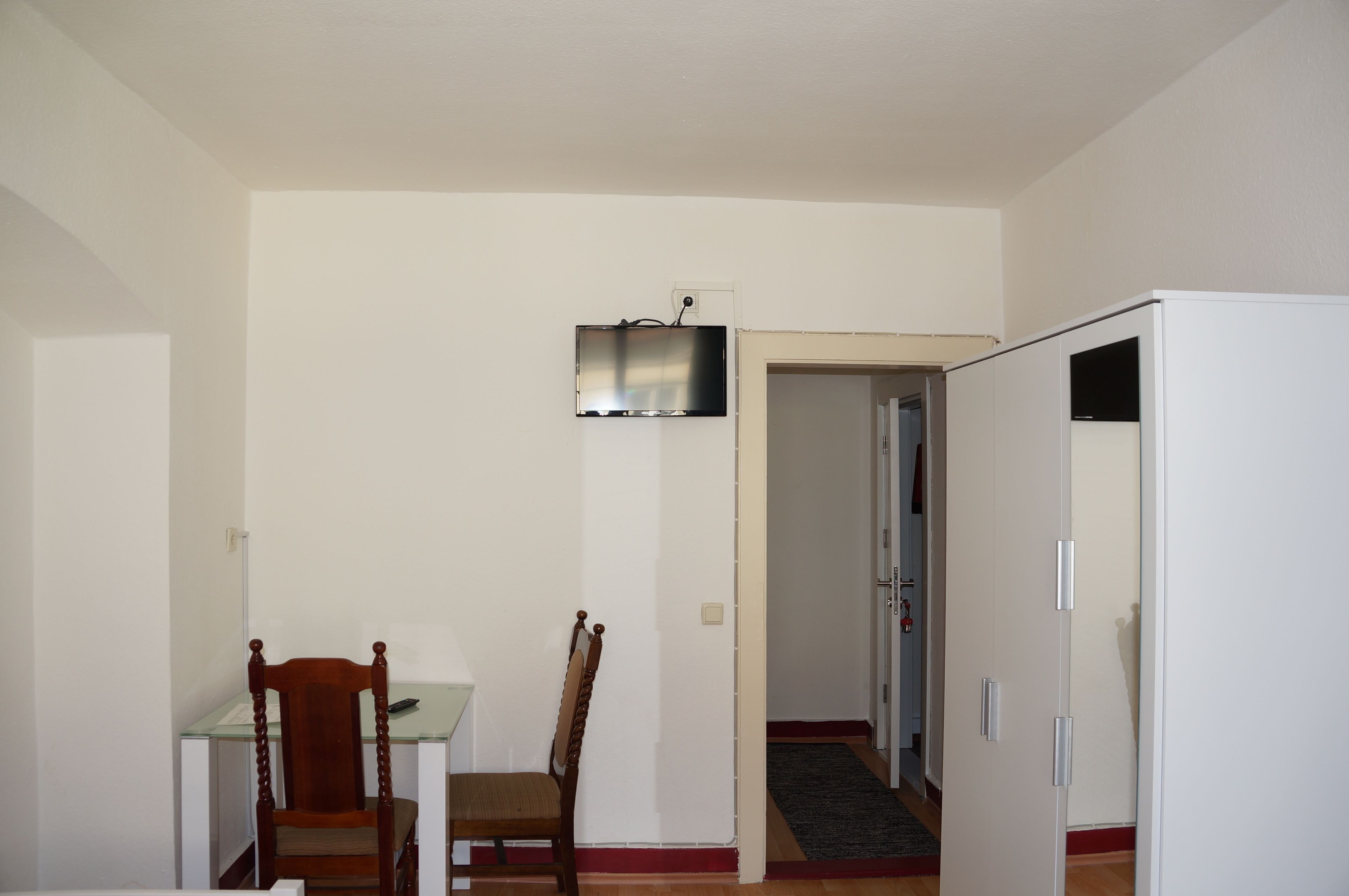 Ein Zimmer mit Esstisch, zwei Stühlen, einem flachbildfernseher an der Wand und einem weißen Kleiderschrank.