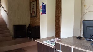 A reception desk with a magazine, a computer, and a chair. There is a staircase in the background and a wall with a framed picture and a fire safety sign. - Eddy Vic Hotel (Abuja)
