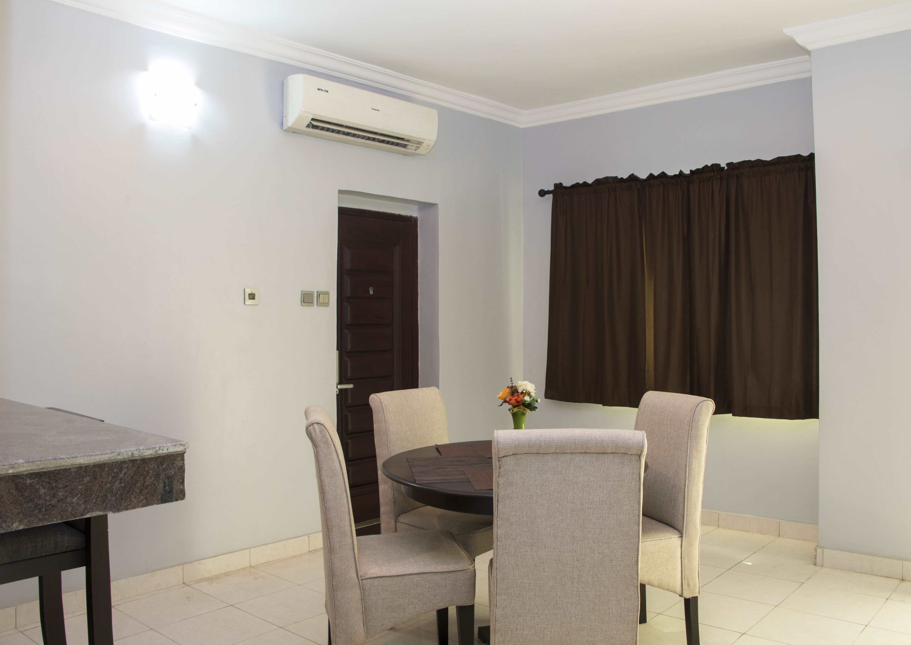 A dining area with a round table, four chairs, an air conditioner, a door, and a window with curtains.