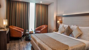 A hotel room with a large bed, two leather armchairs, a small table with a lamp, and a view of the outdoors through the window. - Hide Away Suites (Noida)