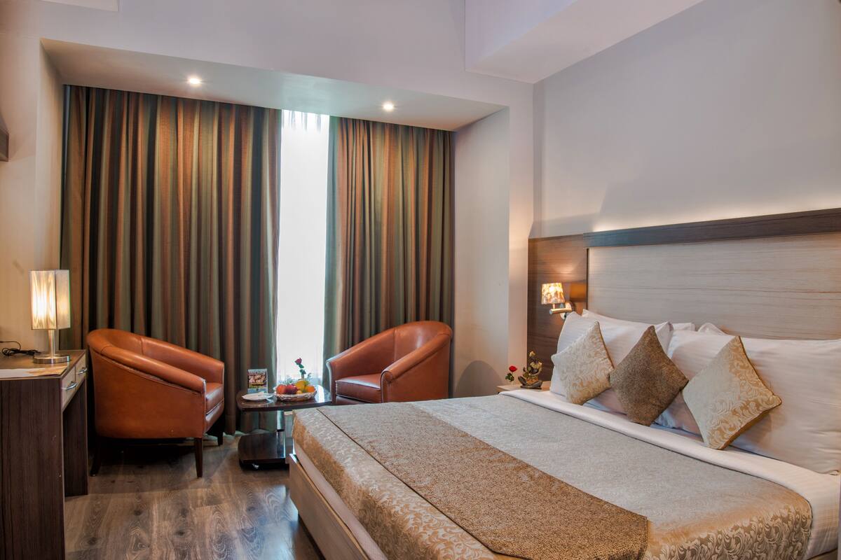 A hotel room with a large bed, two leather armchairs, a small table with a lamp, and a view of the outdoors through the window.