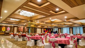 A banquet hall with multiple round tables covered in pink tablecloths, white chairs with pink sashes, and wooden ceiling beams. - Hide Away Suites (Noida)