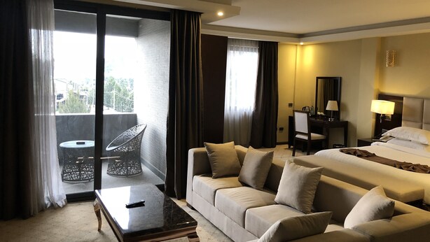 A hotel room with a sofa, a bed, a desk, and a chair. There is a window with curtains and a view of the outside. - Berlottue Hotel (Addis Ababa)