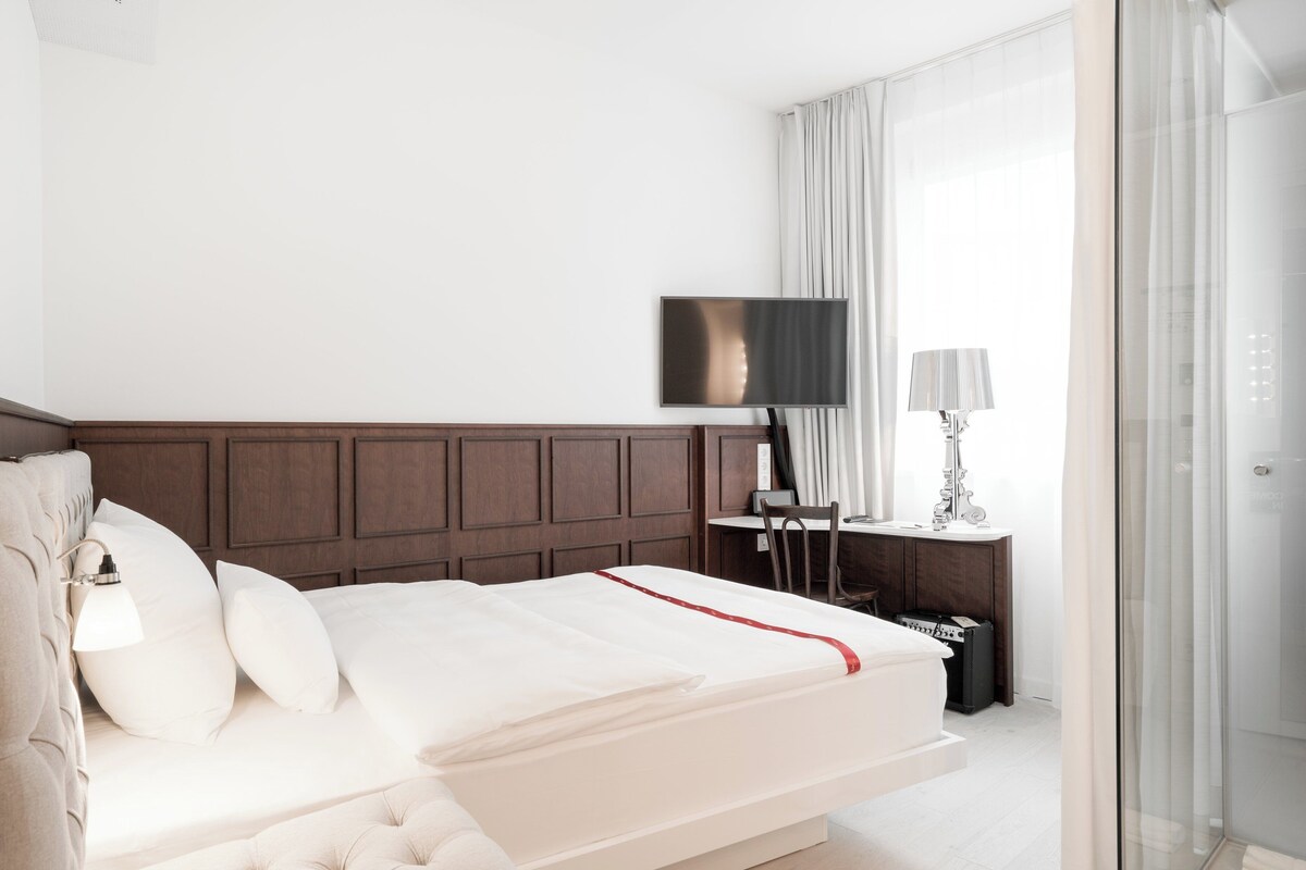 A modern hotel room with a large bed, a flat-screen TV, a bedside table with a lamp, and a glass shower enclosure.