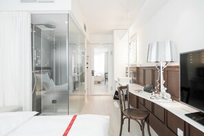 A hotel room with a glass-enclosed shower, a bed with a red accent, a desk with a laptop, a television, and a modern lamp. - Ruby Lissi Hotel Vienna by IHG (Vienna)