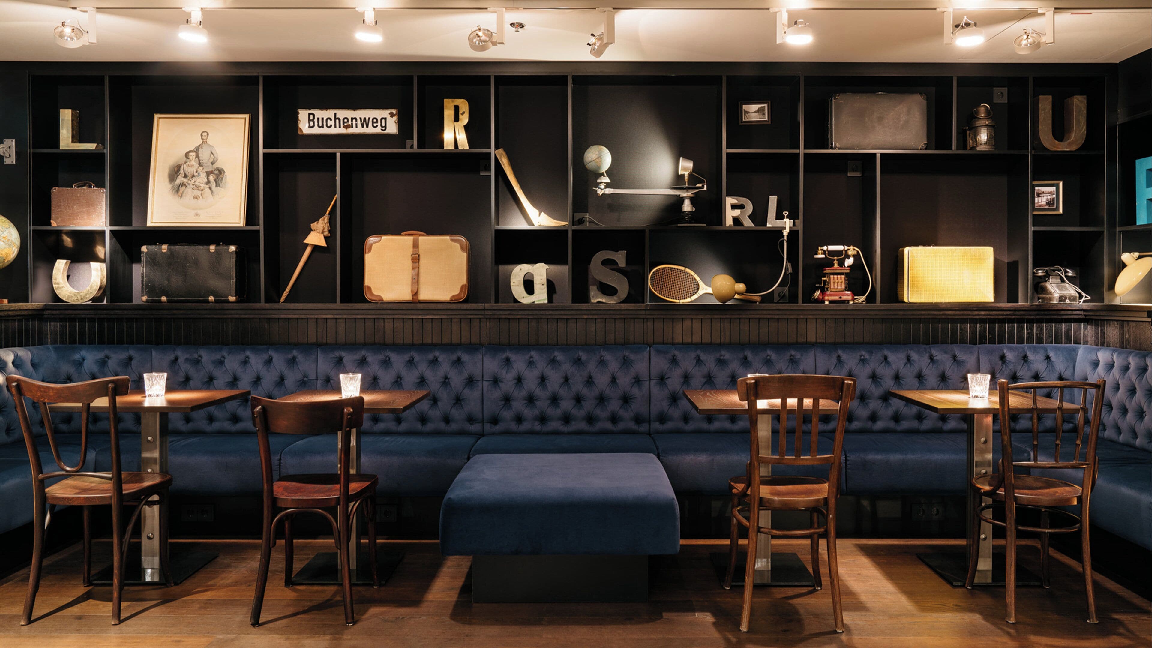 A bar area with a dark blue upholstered bench, wooden chairs, and tables. Shelves above display various items including a globe, a suitcase, and framed pictures.