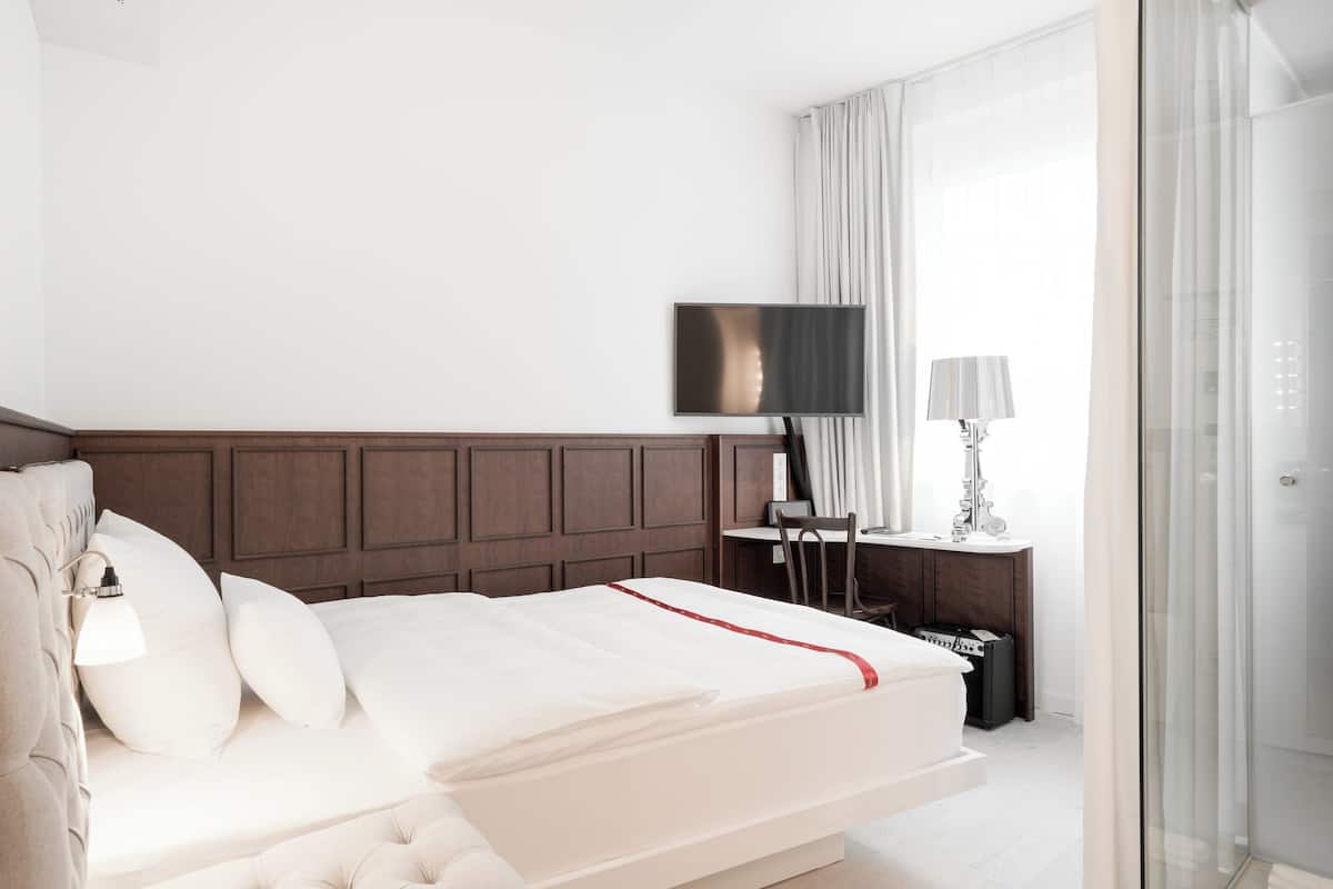 A modern hotel room with a large bed, a flat-screen TV, a bedside table with a lamp, and a glass shower enclosure.