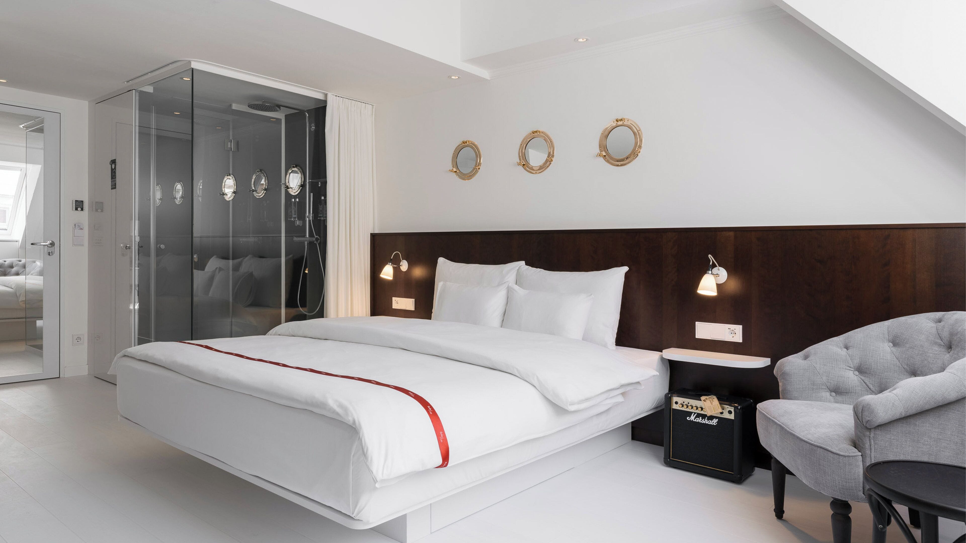 A modern hotel room with a large bed, a glass-enclosed shower, a wooden headboard, and a seating area with a chair and table.