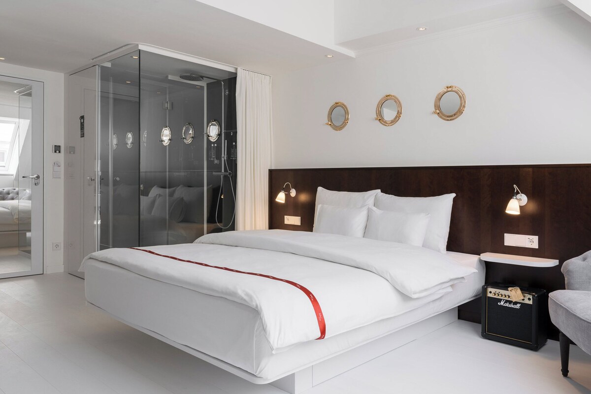 A modern hotel room with a large bed, a glass-enclosed shower, a wooden headboard, and a seating area with a chair and table.