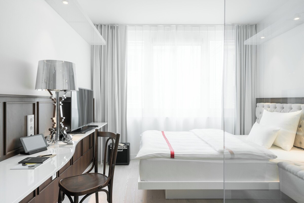 A modern hotel room with a desk, chair, bed, and a large window with sheer curtains.