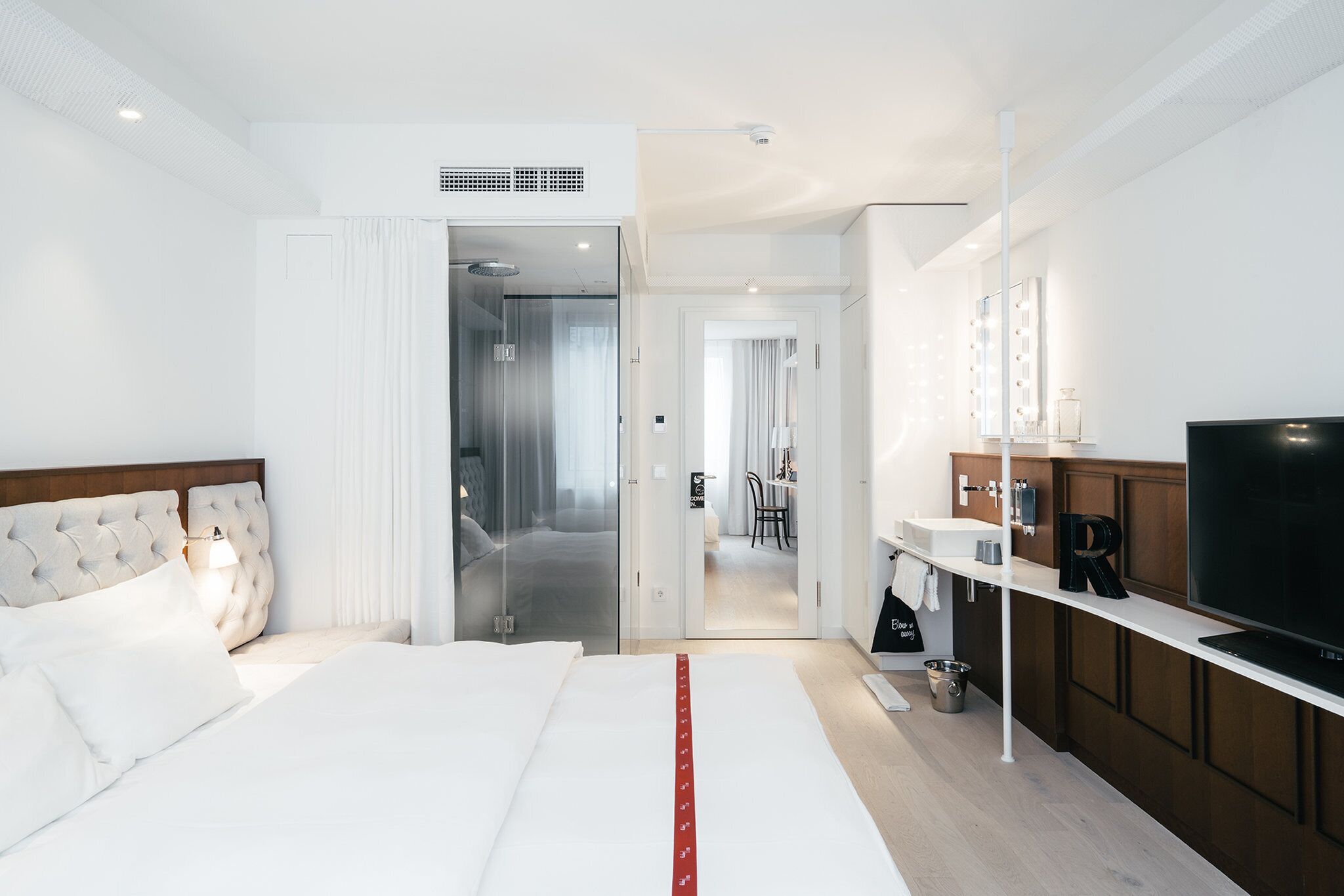 A modern hotel room with a large bed, a TV, a bathroom with a mirror and lights, and a kitchenette.