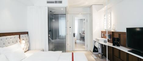 A modern hotel room with a large bed, a TV, a bathroom with a mirror and lights, and a kitchenette.