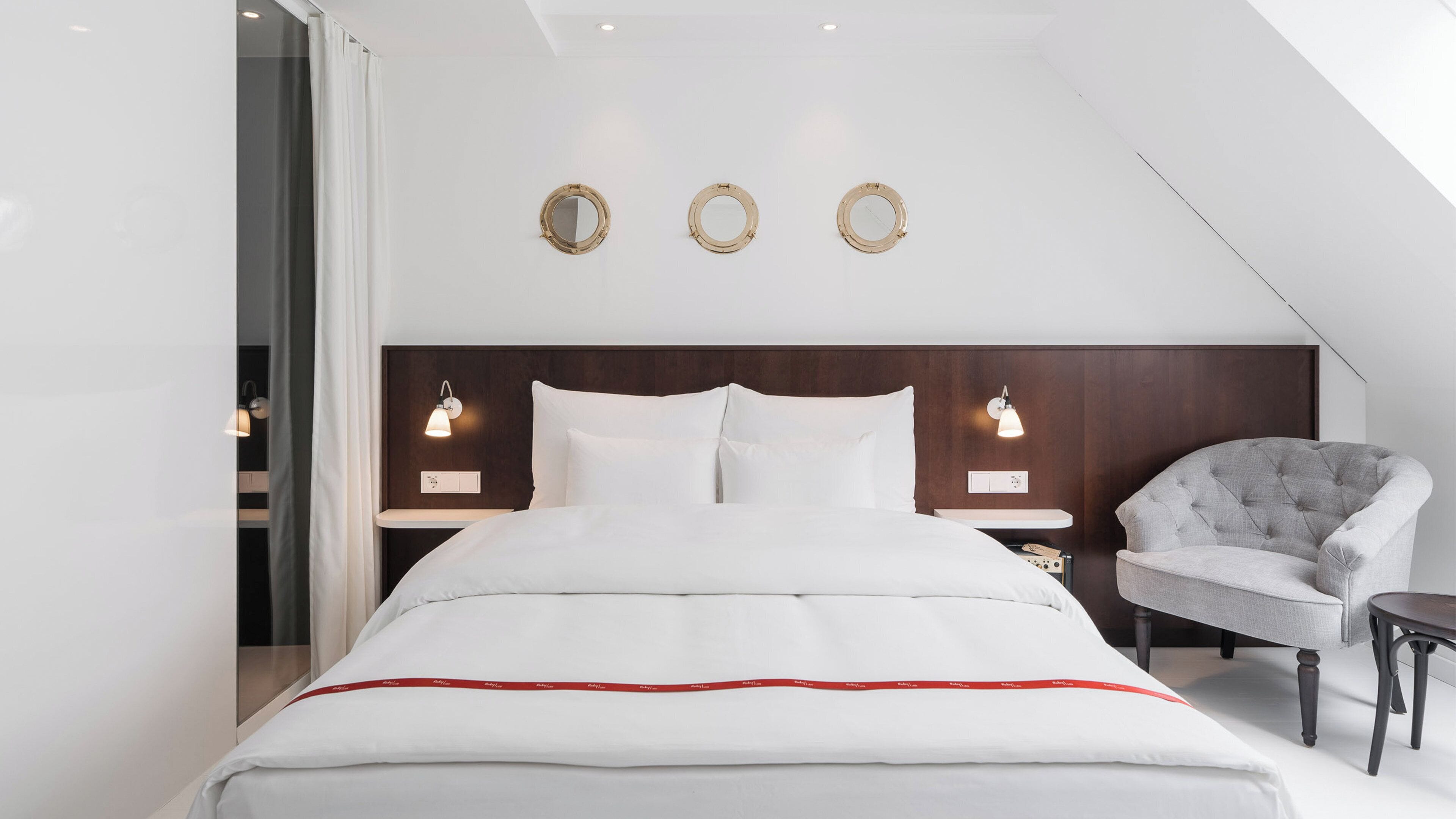A modern bedroom with a large bed, two bedside tables with lamps, a grey armchair, and a small table.