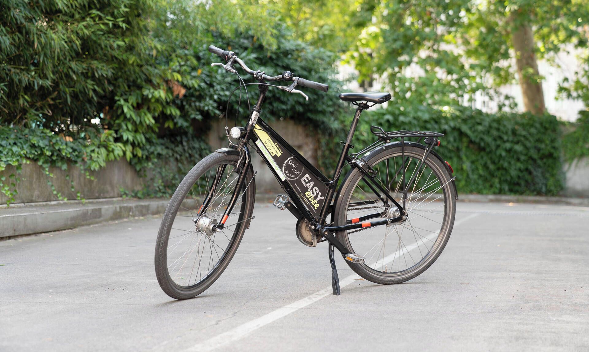 En stillastående cykel med sort stel og et mærke, placeret på en belagt overflade med grøn vegetation i baggrunden.